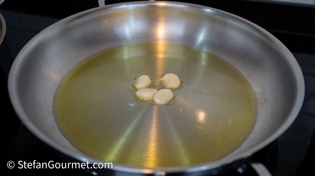A frying pan with hot oil and several cloves of garlic submerged in it.
