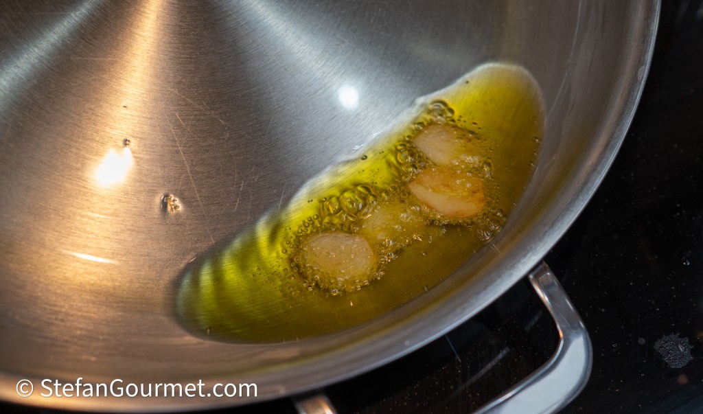 A stainless steel pan with olive oil heating, featuring garlic cloves sizzling in the oil.