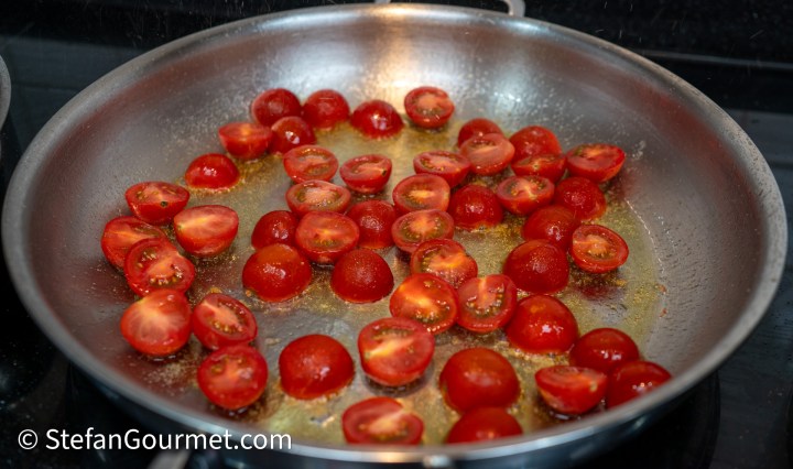 A close-up view of halved cherry tomatoes sizzling in a stainless steel frying pan with olive oil.