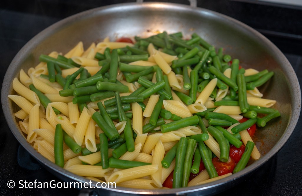 A skillet with cooked penne pasta and fresh green beans mixed together, with some red tomatoes visible in the background.