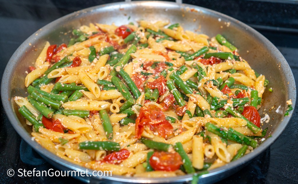 A close-up view of a stainless steel pan filled with a vibrant pasta dish featuring penne, green beans, and cherry tomatoes, all lightly coated in a flavorful sauce.