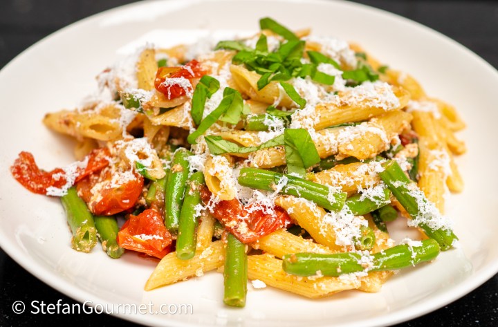 A plate of penne pasta mixed with green beans, cherry tomatoes, topped with fresh basil and grated cheese.
