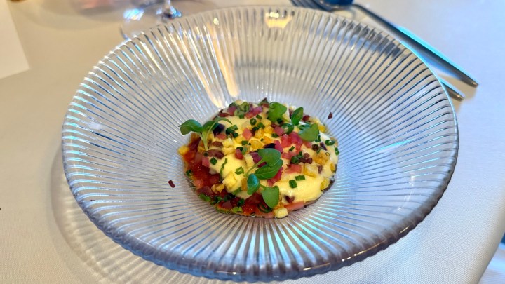 A close-up of a gourmet dish in a clear, ribbed bowl, featuring a creamy base topped with colorful diced vegetables, herbs, and a sprinkle of chives.