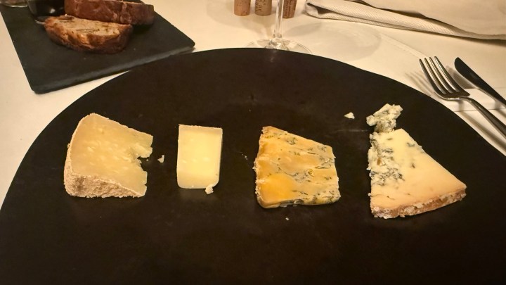 A black plate featuring four different types of cheese arranged in a row, with slices of bread in the background.