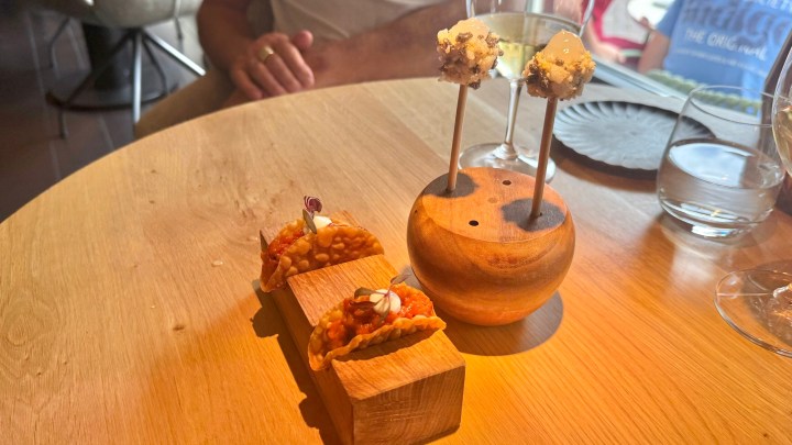 A wooden platter with two decorative mini tacos topped with sauces and garnishes, and a unique wooden stand holding two bite-sized appetizers on skewers, served on a wooden table.
