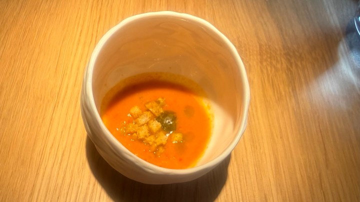 A small bowl containing orange soup with croutons and garnishes, set on a wooden surface.