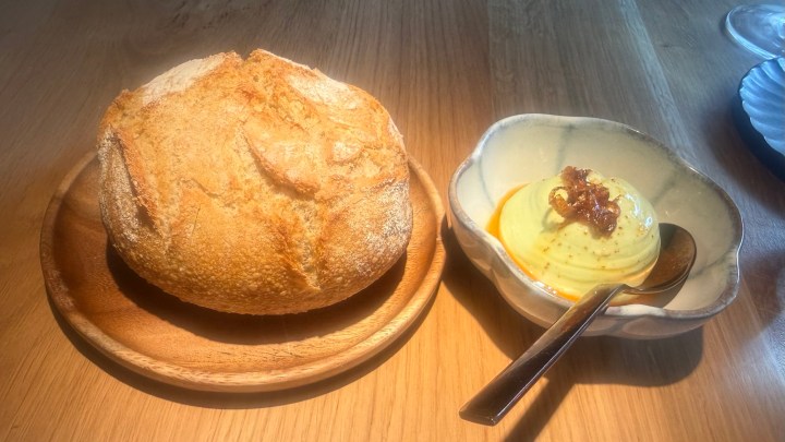 A round loaf of freshly baked bread on a wooden plate next to a bowl of creamy butter topped with a sprinkle of crispy bits, accompanied by a silver spoon.