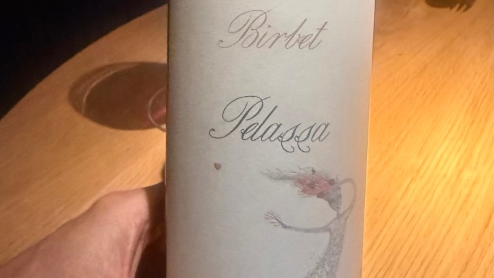 A hand holding a wine bottle labeled 'Birket Pelassa' with a decorative illustration on a wooden table.