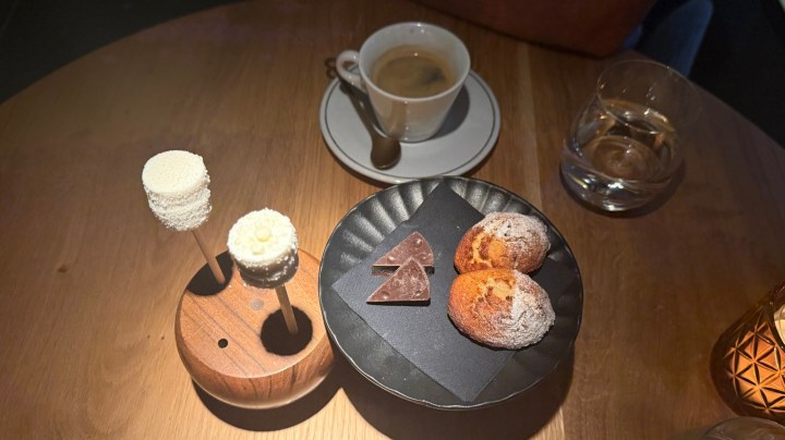 A wooden platter displaying two cake pops, a piece of chocolate, and two small pastries, with a cup of espresso and a glass of water in the background, on a wooden table.