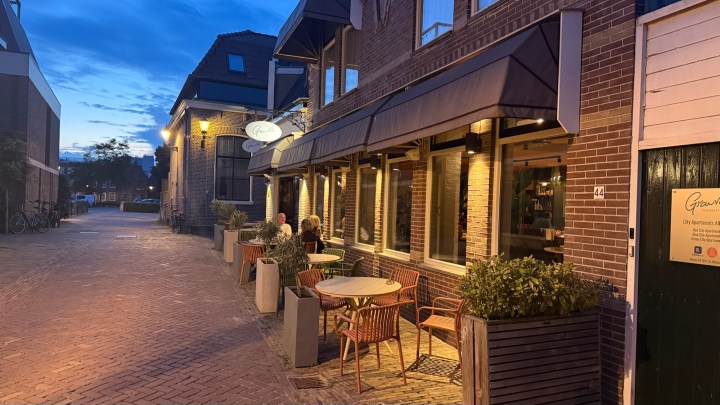 A cozy outdoor dining area with tables and chairs in front of a restaurant at dusk, with warm lights illuminating the space. Two patrons are seated at a table, and brick pavement runs along the street beside the building.