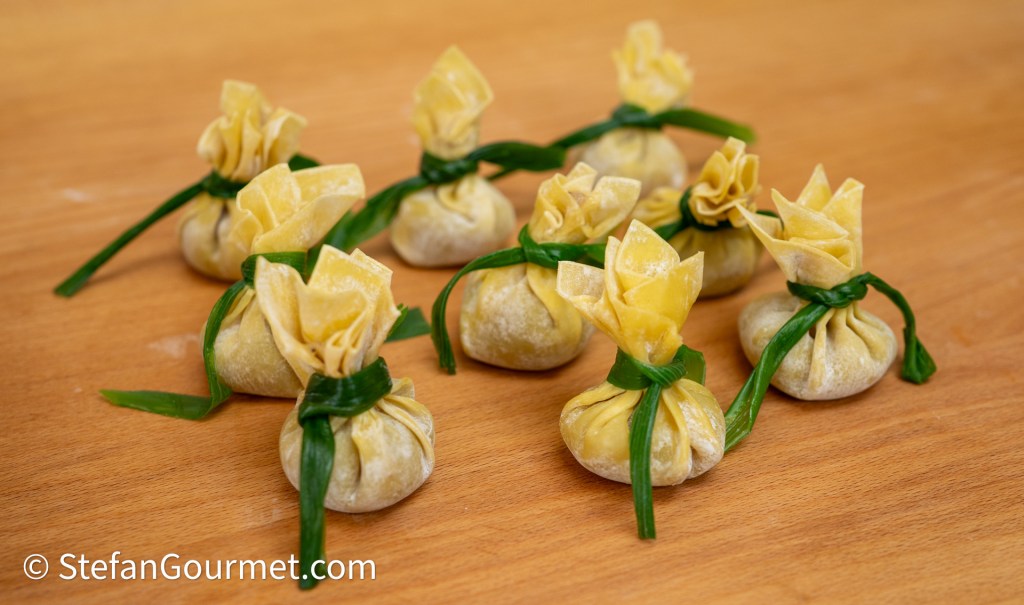 A group of neatly arranged dumplings with yellow wrappers tied with green herbs, placed on a wooden table.