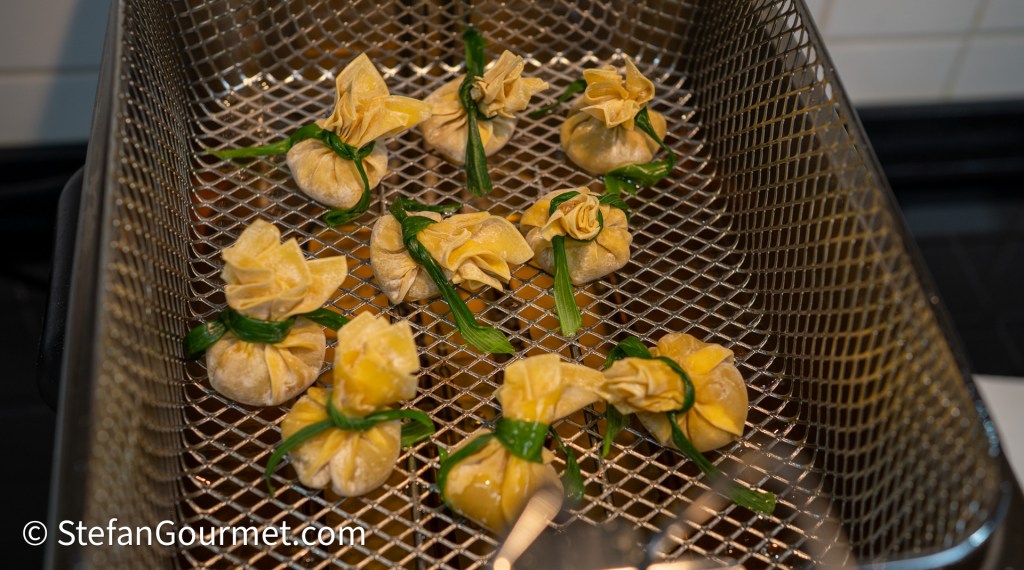 A close-up of dumplings shaped like small pouches, tied with green chives, arranged in a metal steaming basket.