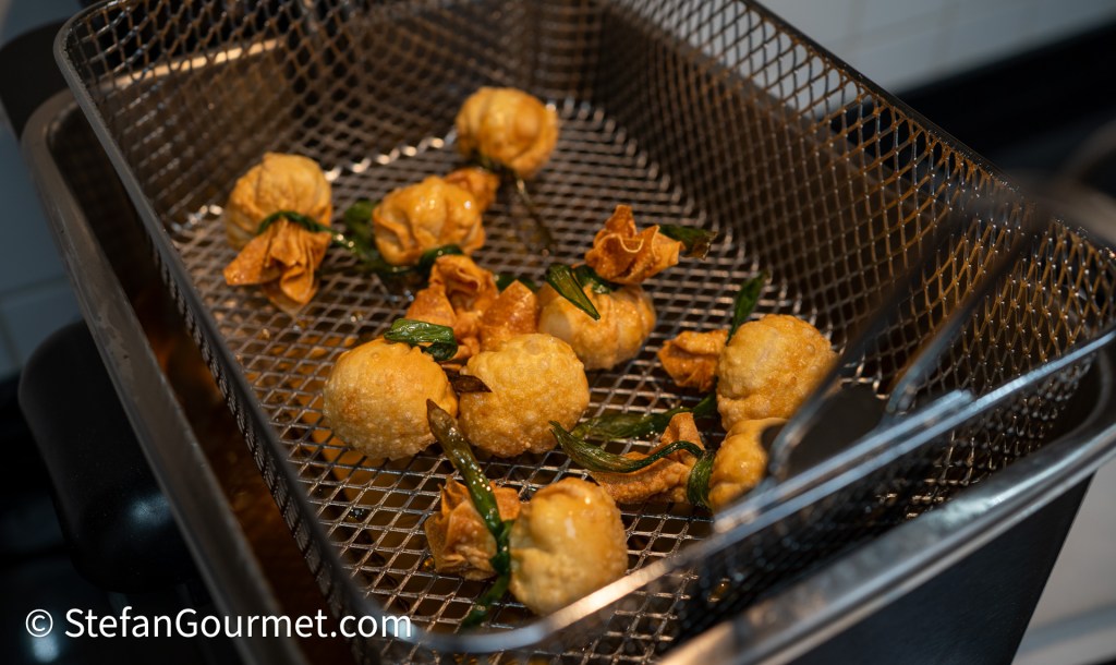 A close-up view of a frying basket containing crispy golden-brown dumplings and crunchy fried wontons, garnished with green herbs.