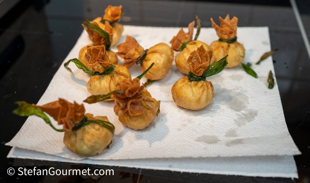A plate of freshly fried dumplings shaped like small bags, garnished with green herbs and crispy pastry, resting on a paper towel.