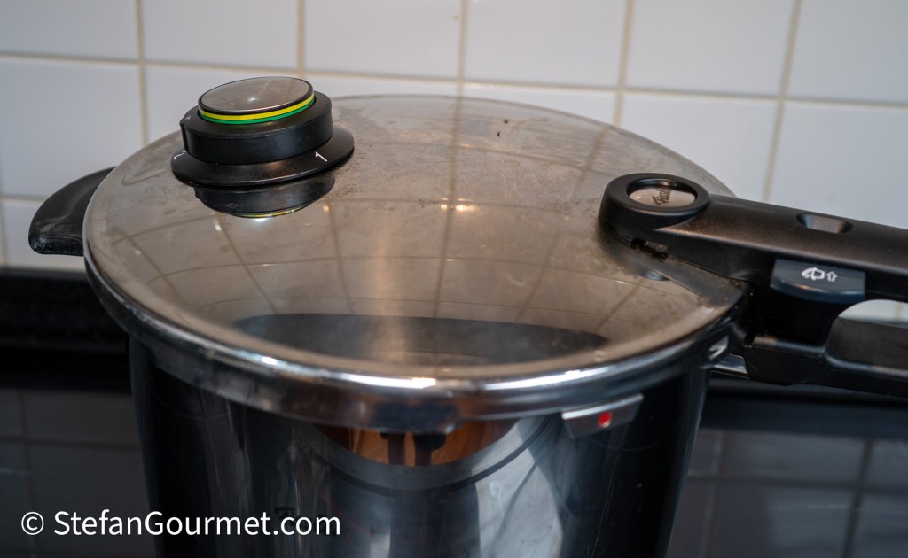 Close-up of a pressure cooker lid with a pressure gauge, reflecting light, set against a tiled kitchen background.