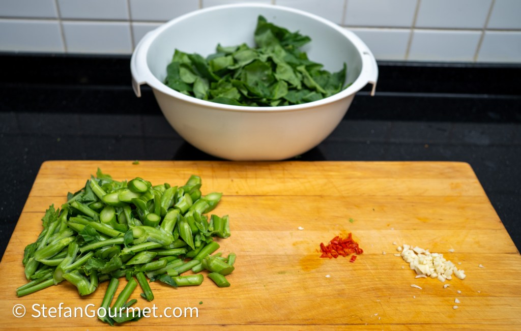 Chopped green vegetables including celery on a wooden cutting board, with minced garlic and red chili flakes, and a bowl of leafy greens in the background.